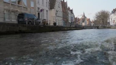 Brugge, Belçika - 2 Nisan 2019: İlkbaharda Brugge'de hızlı hareket eden bir motorlu bottan gelen turistlerin yürüyüşünün olduğu Ortaçağ binalarının etkileyici manzarası ve taş kanal kıyısı