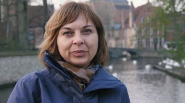 Slo-mo zarif ceket ayakta gülümseyen sarışın bir kadının iyimser portre brugge güzel kuğu ile bir kanal kıyısında duran neşeli kadın ayakta ve slo-mo Brugge bir kanal kıyısında yaptığı gezi hakkında düşünme