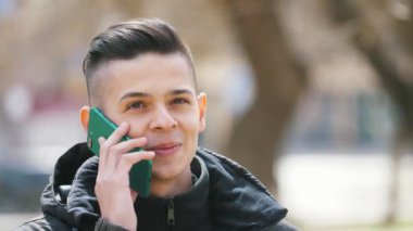 Drolling genç adam akıllı telefon sohbet ve baharda bir sokakta gülümseyen siyah ceketli akıllı bir genç adamın neşeli görünümü gülümseyerek, jesting, ve ilkbaharda parkta onun kız ile akıllı telefon konuşuyor