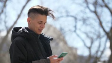 Komik genç adam ayakta ve ilkbaharda onun akıllı telefon açık havada arama Jolly bokeh siyah ceketli mutlu bir genç adam görünümü onun akıllı telefon net sörf ve baharda bir sokakta neşeyle gülümseyen
