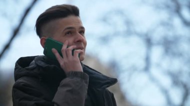 Gülümseyen genç brunet adam baharda bir sokakta akıllı telefon sohbet Siyah ceketli akıllı bir genç adamın neşeli görünümü gülümseyerek, ayakta ve ilkbaharda bir parkta mutlu bir şekilde akıllı telefon üzerinde konuşurken