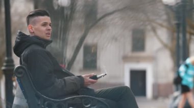 Snappy brunet adam oturma ve ilkbaharda bir parkta bir parkta e-sigara sigara açık havada bir bankta oturan ve ilkbaharda sokakta elektronik sigara içen siyah bir ceket kısa bir saç kesimi ile genç bir adam etkileyici görünümü