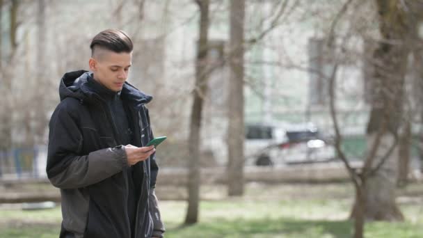 在春天的公园里 一个穿着黑色夹克的年轻布鲁内特男子在智能手机上浏览网 在春天的小巷里愉快地微笑着 在春天的公园里 一个活泼的年轻人站在一个公园里 用智能手机站在智能手机上 用智能手机拨通自己的智能手机 — 图库视频影像
