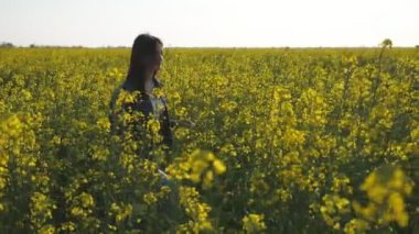 Muhteşem kız sarı bir kolza alanında geziniyor ve slo-mo bir kot takım elbise li uzun gevşek saçlı mutlu bir esmer kız neşeli görünümünde gülümsüyor slo-mo yaz aylarında bir çiçek li kolza alanında yürüyüş