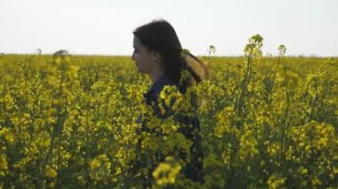 Güzel kız bir çiçekli kolza alanında gidiyor ve slo-mo bir kot takım elbise uzun gevşek saçlı neşeli bir esmer kız slo-mo muhteşem profili gülümsüyor slo-mo yaz aylarında gelişen bir kolza alanında gezinen