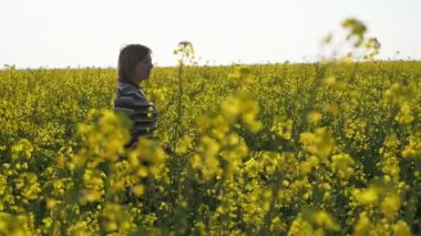 Mutlu kadın gidiyor ve slo-mo bir alanda kolza çiçekleri bakarak çizgili bir bluz ve kot yürüyen gülümseyen sarışın bir kadının harika profili yürüyüş ve slo-mo bir alanda sarı kolza çiçekleri görünümlü