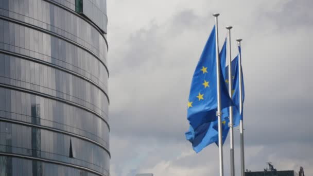 曇りの日に欧州議会で揺れる3つのEu旗は スロモの春にガラスとコンクリートで作られた欧州議会の建物で栄光に振るいくつかのEuの旗の印象的な眺め — ストック動画