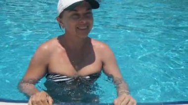 Slo-mo'da bir yüzme havuzunda ayakta ve gülümseyen neşeli kadın, türkiye'de yavaş çekimde kürek havuzunda duran beyzbol şapkası ve mayo içinde mutlu bir kadın turistin neşeli görünümü