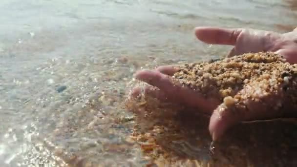 スローモーションで夏の晴れた日に黒海の海岸で砂 輝く水を保持する女性の手の感情的なビューで海岸で砂と水で遊ぶ女性の手 — ストック動画