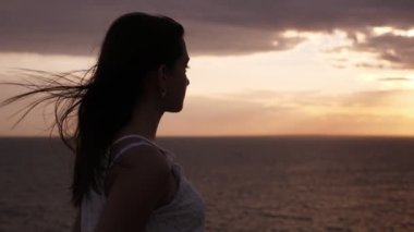 Slo-Mo karanlık günbatımı bir sallayarak denize bakarak beyaz bir elbise düşünceli kız bir deniz plajında ayakta uzun saçlı çekici esmer kız Ilham verici görünümü ve Slo-Mo yaz aylarında karanlık günbatımı köpüklü deniz zevk