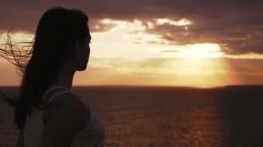 Slo-Mo karanlık günbatımı bir dalgalanan denize bakarak beyaz bir elbise içinde Dreamy kız bir deniz kıyısında ayakta uzun saçlı güzel bir esmer kız romantik görünümü ve yavaş hareket yaz aylarında karanlık gün batımı içinde sallayarak deniz zevk