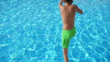 Sarı şortlu komik çocuk, yavaş hareket içinde bir Wading gölet ilk ayaklar atlama sarı şort mutlu bir Brunet çocuk güzel bir yüzme gölet içinde parlayan turkuaz suları ile ilk atlama ayaklar.