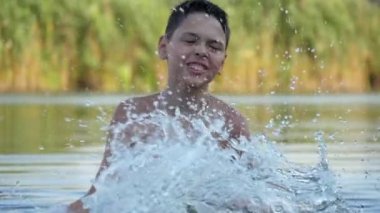 Komik küçük çocuk ayakta ve kısa saç kesimi ayakta neşeli küçük brunet çocuk neşeli küçük brunet çocuk duygusal görünümünde göl suyu sıçrama elleri dönen, onun hareketli elleri ile su sıçraması ve slo-mo yaz aylarında bir kamış gölünde eğlenceli