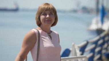 Neşeli sarışın kadın ayakta ve slo-mo yaz aylarında Dnipro nehir de gülümseyen bir deri çanta ayakta ve slo-mo yaz güneşli bir günde Dnipro nehir de neşeli gülümseyen açık pembe elbiseli ince bir sarışın kadın harika görünümü