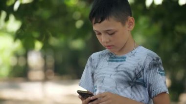 Küçük çocuk ayakta ve yaz aylarında yeşil bir parkta hızlı video oyunları oynarken renkli bir t-shirt ayakta akıllı küçük brunet çocuk etkileyici görünümü ve yaz güneşli bir günde bir parkta bir profesyonel gibi akıllı telefon videooyunları oynarken