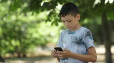 Akıllı esmer çocuk yaz aylarında bir parkta onun smartphone üzerinde video oyunları oynarken bir T-shirt ayakta ve yaz aylarında güneşli bir günde yeşil bir parkta onun smartphone üzerinde video oyunları oynarken neşeli küçük esmer çocuk heyecan verici görünümü 