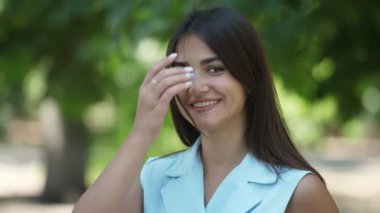 Yaz aylarında yeşil bir parkta sırayla saçlarını koyarak parlayan gözleri ile mutlu kız açık mavi elbise ayakta uzun saçlı neşeli bir esmer öğrencinin harika görünümü ve yaz aylarında güneşli bir gün sokakta mutlu bir gülümseme ile fonksiyonunuz