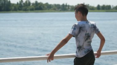 Küçük çocuk ayakta ve tutmak, bir T-shirt ve pantolon ayakta kısa saç kesimi ile neşeli bir esmer çocuk ve Slo-Mo güneşli bir günde Dnipro dalgalanan dalgalar zevk Slo-Mo muhteşem arka görünümünde Dnipro su bakıyor korkuluklar
