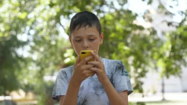 T-shirt akıllı çocuk yavaş hareket ormanda onun telefonda video oyunları oynarken bir T-shirt ayakta ve yavaş hareket yaz aylarında güneşli bir günde yeşil bir ormanda onun akıllı telefonda video oyunları oynarken mutlu bir esmer çocuk harika görünümü