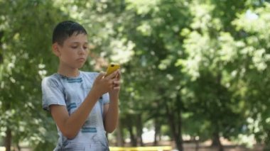Bir T-shirt ayakta ve yavaş hareket yaz aylarında güneşli bir günde bir yeşil parkta onun akıllı telefonda video oyunları oynarken bir akıllı esmer çocuk yavaş hareket Cheery görünümünde bir sokakta telefonda video oyunları oynarken T-shirt mutlu çocuk