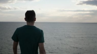 Cheery esmer adam deniz kıyısı kenarına seyir Slo-Mo etrafında siyah deniz sahilinin çimenli kenarına yürüyüş ve SLO-MO içinde duygusal yaz etrafında görünümlü çırpınan saç ile ince bir genç adam harika arka görünüm gidiyor