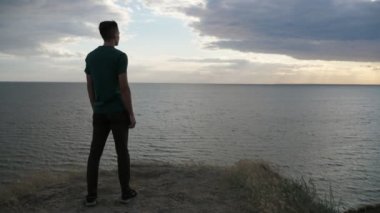 Sportive genç adam ayakta ve Slo-Mo çarpıcı zoom-arka görünümünde bir T-shirt ve siyah pantolon ayakta ve güçlü Karadeniz 'de yaz aylarında çırpınan saç ile atletik bir genç adam içinde Black Sea görünümü zevk-Mo