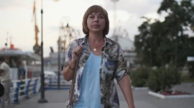 Kendinden emin ve şık sarışın kadın slo-mo yaz aylarında Dnipro rıhtım üzerinde gezinen bir deri çanta ile modish renkli gömlek, T-shirt, bob saç kesimi ile başarılı bir sarışın kadın başarılı bir sarışın kadın yaz aylarında Dnipro rıhtım üzerinde yürüyüş  