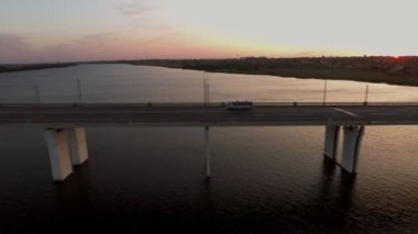 Yaz aylarında gün batımında Ukrayna'da hareket eden bir otobüs ile uzun bir köprünün havadan çekilen hareketli bir otobüs ve yaz aylarında gün batımında aynı hızda bir kenara uçan bir drone ile Dnipro nehri üzerinde uzun bir modern köprü şaşırtıcı kuş bakışı görünümü.
