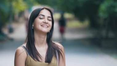 Uzun saçlı neşeli brunet kız gülümseyerek ve mutlu yeşil bir sokakta gülüyor Uzun gevşek saçlı ayakta güzel bir brunet kız Amazing bokeh portre, gülümseyerek ve yaz aylarında güneşli bir günde yeşil bir sokakta mutlu bir şekilde gülüyor.