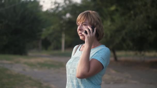 夏の緑の公園で電話で話す美しいブロンドの女性は スローモーションで夏の晴れた日に緑の公園で立って 彼女のスマートフォンでおしゃべりしているスマートなブロンドの女性のスロモム楽観的なビューで — ストック動画