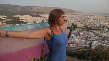 Tepede duran ve Atina 'da arkasından pareosunu geren neşeli bir kadın. Bir tepede dikilen ve sırtında şeffaf pareosuyla Atina manzarasının tadını çıkaran mutlu sarışın bir kadın manzarası heyecan verici..