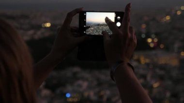 Telefon tutan kadın parmaklar ve Atina şehir manzarasının gece çekilmiş bir fotoğrafı modern bir akıllı telefonu elinde tutan ve yazın karanlık gökyüzü ile Atina şehir manzarasının fotoğrafını çeken kadınların etkileyici bir yakın çekimi. Harika görünüyor..