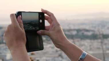 Akıllı telefon tutan ve Atina manzarasının fotoğrafını çeken kadın elleri modern bir akıllı telefonu kavrayıp güneşli bir yaz gününde sınırsız ufuk çizgisi olan Atina manzarasının fotoğrafını çekiyor..