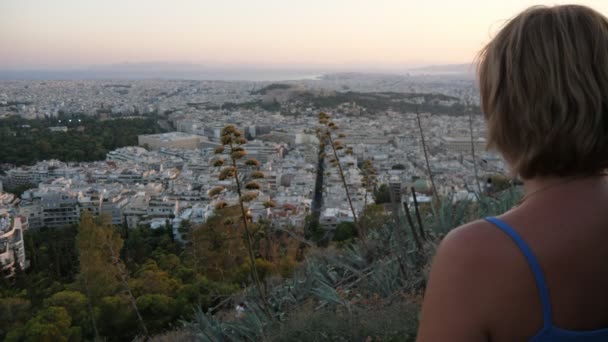 丘の上に立つスレンダーブロンドの女性と夏にアテネの景色を見て印象的なバック夏には古代都市の街並みを見て 丘の上に立って無袖ブラウスにボブヘアカットと好奇心ブロンドの女性のビュー — ストック動画