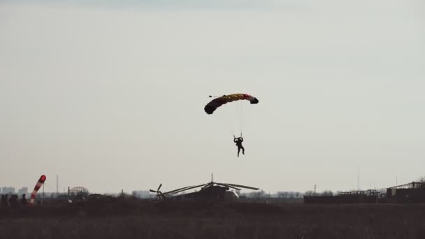 军用跳伞员在秋日的崎岖田野上降落在方降落伞上 一名身着军装的军人在秋日的傍晚降落在崎岖的草地上 直升机被看到很远 — 图库视频影像