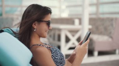 Neşeli esmer kız bir şezlong üzerinde yatan ve yaz aylarında telefonunda net tarama Bir şezlong ve güneş gözlüğü uzun gevşek saçlı gülümseyen esmer kız profili bir şezlong üzerinde yatan ve bir deniz beldesinde onu telefonda net sörf