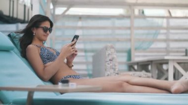 İnce esmer kız bir şezlong üzerinde yatan ve yaz aylarında telefonunda net sörf Bir şezlong ve güneş gözlüğü bir şezlong üzerinde yatan ve bir deniz beldesinde onu telefonda net tarama uzun gevşek saçlı şık bir esmer kız güzel görünümü