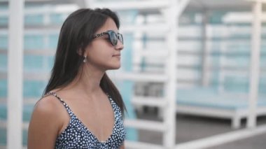 Bikini ayakta neşeli kız ve yaz aylarında güneşlikler kadar smartening Bikini ve güneş gözlüğü ayakta uzun gevşek saçları ile mutlu bir genç brunet kadının harika görünümü, gülümseyen ve bir deniz beldesinde mavi güneşlikler kadar smartening