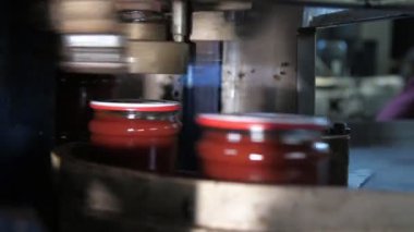 Domates ezmesi kavanozlarına çarpan bir domates ezmesi kavanozunun içindeki yuvarlanan bir robotun altında domates işleme fabrikasında, domates hamuru altında metal bir şeritte hareket eden domates hamuru bantlarının kapaklarını kapatıyor. Neşeli görünüyor..