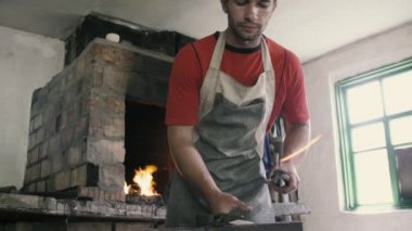 Profesyonel demirci tuğladan kırmızı çubuğu ateşle alıyor. Pamuk önlüklü genç bir demircinin forsepsli metal kavisli çubuğu alıp, örsü takıp atölyenin içine çekle vuruşunu görüyor..