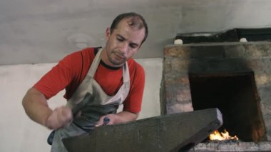 Demirci, eski bir atölyedeki çekiçle metalik bir gülü vuruyor. Pamuk önlüklü bir demircinin sıcak metalik bir gülü çekiçle vuruşunu ve eski bir atölyede, tuğla fırınıyla döndürüşünü hayrete düşürüyor..