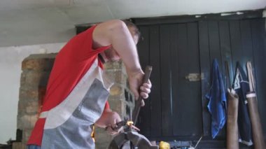 Yetenekli demirci eski bir atölyede çekiçle kırmızı kamaraya vuruyor. Pamuk önlüklü bir demircinin çekici ile sıcak kavisli bir oltaya vurduğunu ve eski bir atölyede tuğla fırınla daire çizdiğini görüyor..