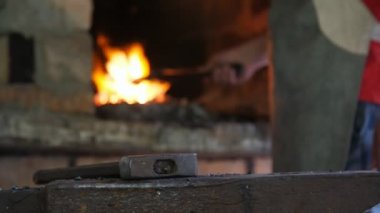 Erkek el, bir atölyedeki maşayla fırından metal bir kaleyi alıyor. Erkek eli, fırından maşayla kırmızı metalik bir kaleyi alıyor ve eski bir demirci atölyesinde çekle örse takıyor. 