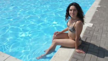 Güneşli bir günde kürek çeken bir gölette gülümseyen, bikinili, uzun kıvırcık saçlı, uzun saçlı, güneşli bir yaz gününde gölet kaldırımında bacak bacak üstüne atmış bir kadının harika manzarası..