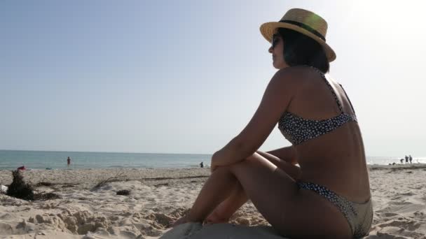 穿着比基尼的时尚女人坐在海滩上 听着音乐 看到一个身材苗条 戴着太阳镜和草帽的年轻女子坐在那里 玩沙子 夏天在黑海海滩听音乐 — 图库视频影像