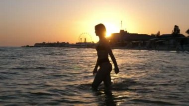 Cazibeli kız gün batımında şehir rıhtımında ışıl ışıl nehir sularına giriyor. Çekici, kısa saçlı, bikinili esmer bir kızın altın gün batımında dönme dolap ile şehir setinde parlayan nehir dalgalarıyla giderken görüntüsü ilham verici.