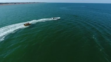 Yazın açık denizde mutlu turistlerle muz çeken güçlü bir teknenin havadan çekilmiş görüntüsü güneşli bir yaz gününde komik insanlarla birlikte şişme muz çeken büyük beyaz bir motorlu botun neşeli görüntüsü. Yavaş yavaş sağa dönüyor..
