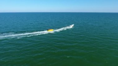 Yazın açık denizde insanlarla muz çeken motorlu teknenin hava görüntüsü güneşli bir yaz gününde neşeli insanlarla dolu şişme bir muzu çeken güçlü beyaz bir motorlu botun iyimser görüntüsü. Adım adım sola dönüyor..