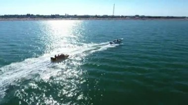 Yazın açık denizde insanlarla birlikte muzları çeken motorlu bir hava aracı. Güçlü beyaz bir motorlu teknenin güneşli bir yaz gününde neşeli insanlarla dolu şişirilebilir bir muzu çekişi. Sağa dönüyor..