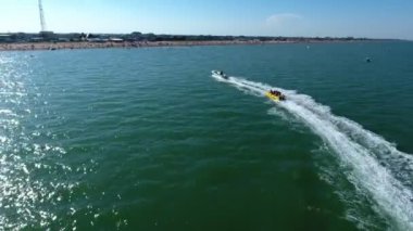 Yazın açık denizde turistlerle birlikte şişme muz çeken motorlu hava aracı. Güneşli bir yaz gününde, güçlü beyaz bir motorbotun şişirilebilir bir muzu, neşeli insanlarla birlikte açık denizde sürüklediği harika bir manzara. Sola dönüyor..